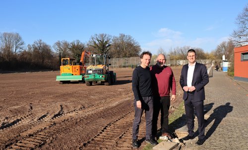 Drei Männer auf einem umgepfügten Feld, dahinter eine Baumaschine