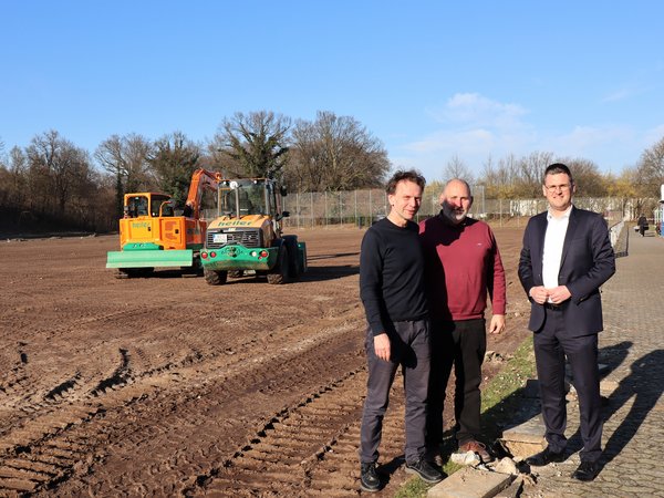 Drei Männer auf einem umgepfügten Feld, dahinter eine Baumaschine