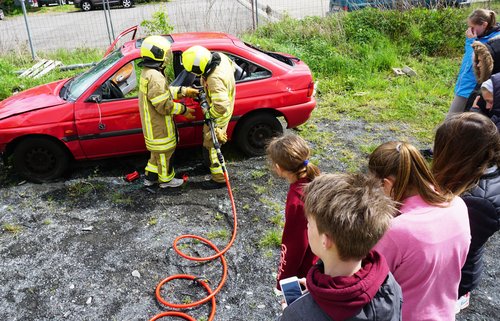 Feuerwehrleute schneiden ein Auto auf, Jugendliche schauen zu