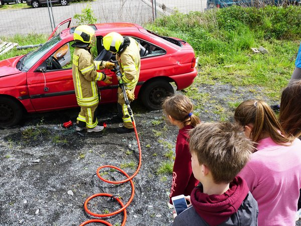 Feuerwehrleute schneiden ein Auto auf, Jugendliche schauen zu