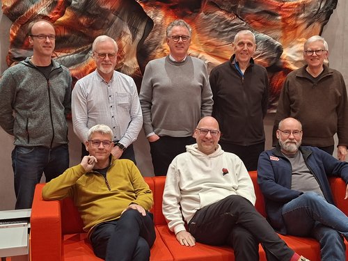 Drei Männer sitzen auf einer roten Couch, fünf stehen dahinter