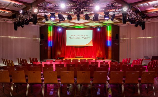 Saal des Freizeithauses, bestuhlt und die Bühne ist farbig beleuchtet. 