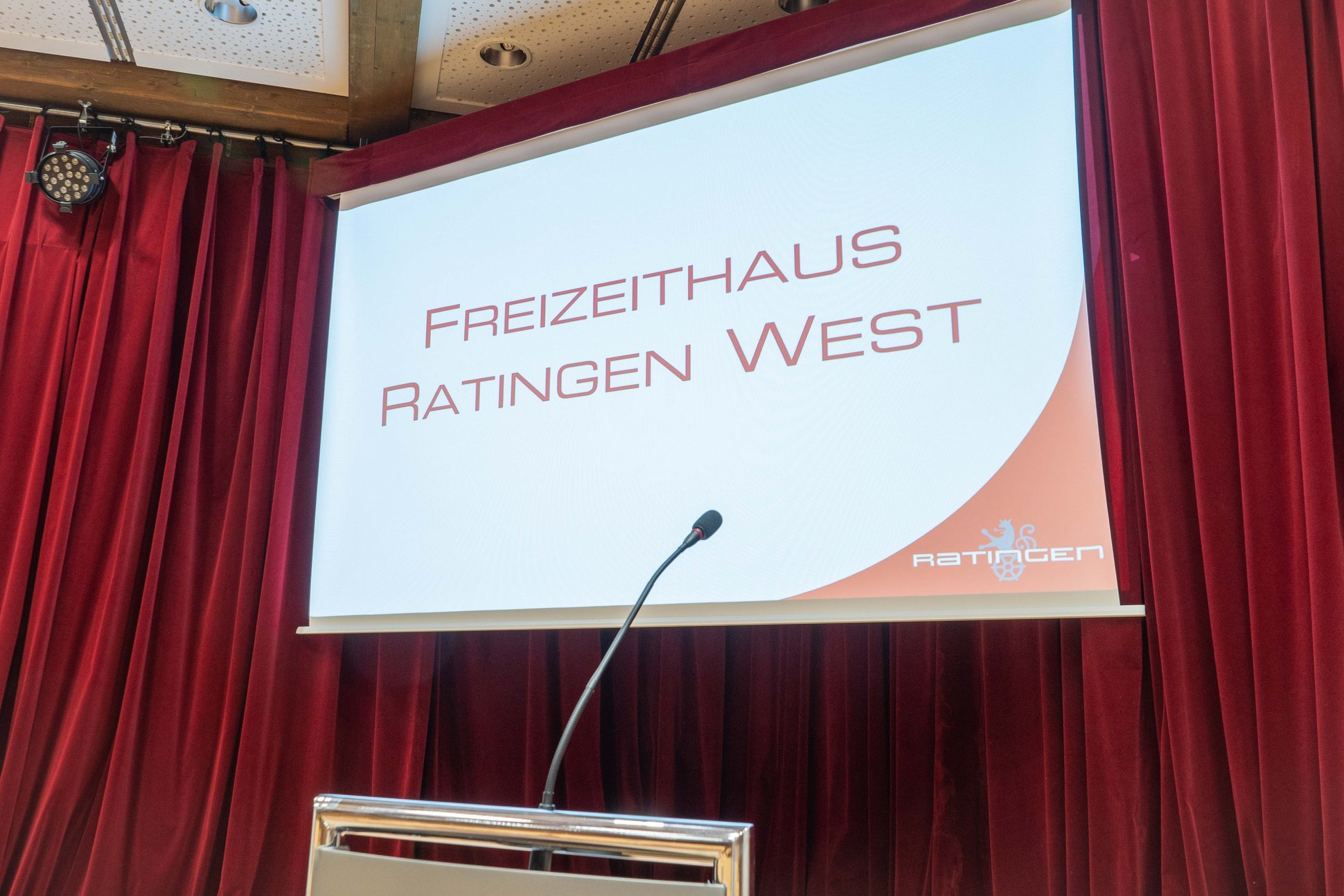 Leinwand auf der Bühne des Saals im Freizeithaus, im Hintergrund rote Vorhänge. 