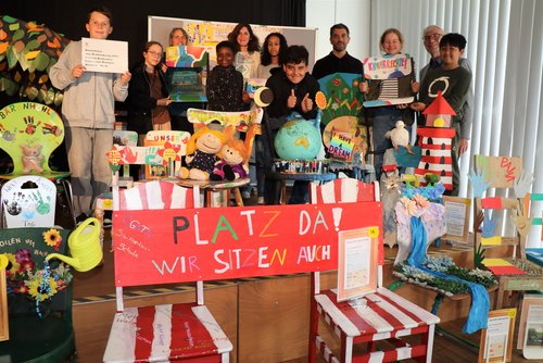 Kinder und weitere Personen auf einer Bühne mit kunstvoll gestalteten Stühlen