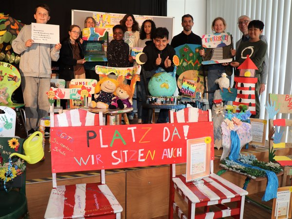 Kinder und weitere Personen auf einer Bühne mit kunstvoll gestalteten Stühlen
