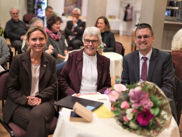 Drei Personen, zwei Frauen und ein Mann, sitzen lächelnd an einem Tisch