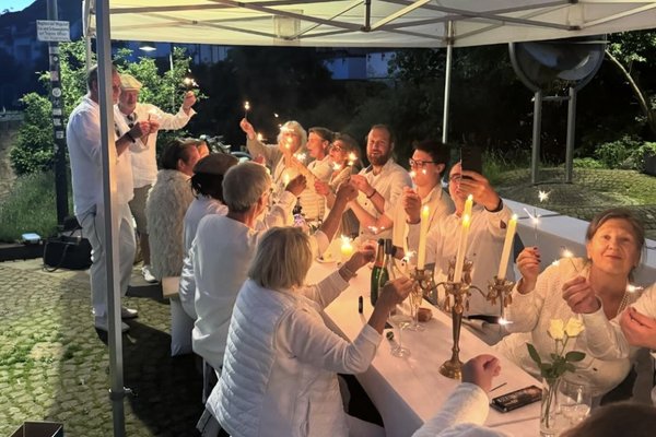 Menschengruppe ganz in weiß gekleidet sitzt am festlich gedeckten Tisch. 