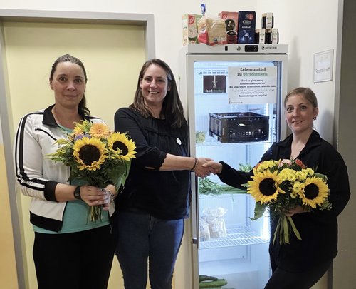 Drei Frauen vor einem Kühlschrank, zwei halten Blumensträuße in den Händen