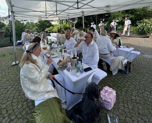 Personen in weiß gekleidet sitzen an Bierzeltganituren, im Hintergrund spielt eine Band