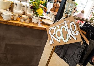 Das Bild zeigt Kaffeegeschirr und einen Kleiderständer, an dem das selbst gemalte Schild "Röcke" hängt.