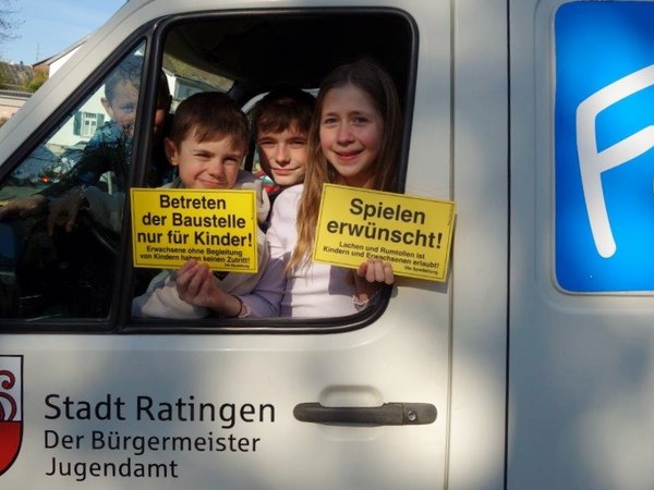 Drei Kinder schauen auf einem Autofenster heraus und halten kleine Schilder in der Hand 