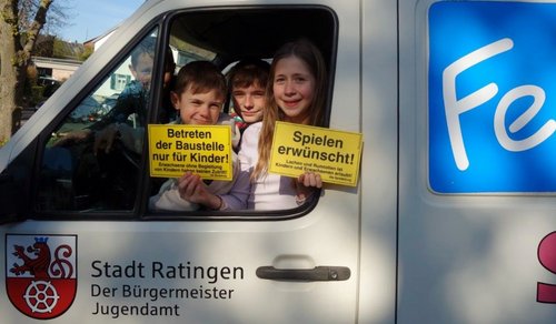 Drei Kinder schauen auf einem Autofenster heraus und halten kleine Schilder in der Hand 