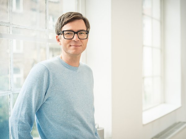 Mann mit braunen Haaren, schwarzer Brille und hellblauem Pullover vor einem großen Fenster.