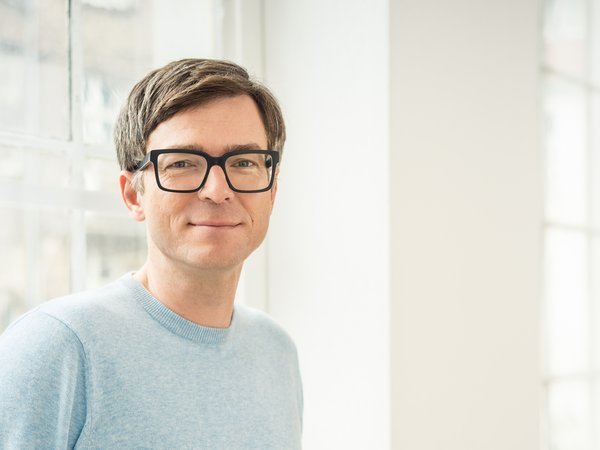 Mann mit braunen Haaren, schwarzer Brille und hellblauem Pullover vor einem großen Fenster.