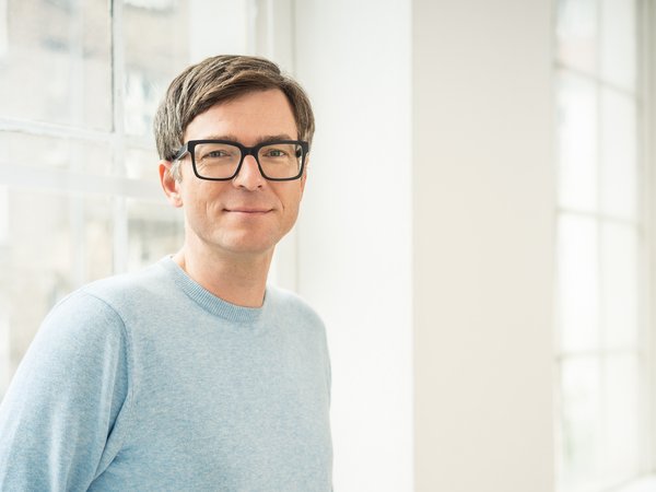 Mann mit braunen Haaren, schwarzer Brille und hellblauem Pullover vor einem großen Fenster.