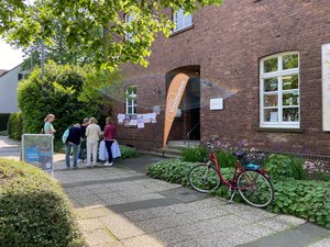 Außenfassade des Weltladen Lintorfs mit einem Infostand, an dem sich Besuchende mit den Mitarbeitenden des Weltladens unterhalten. 