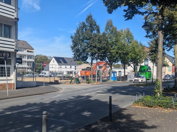 Straße mit Bäumen und Wohnhäusern, gegenüber eine Baustelle mit Bagger und Lastwagen
