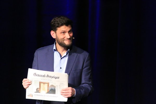 Christian Ehring steht grinsend vor einem dunklen Vorhang und hat die Zeitung "General Anzeiger" in der Hand.