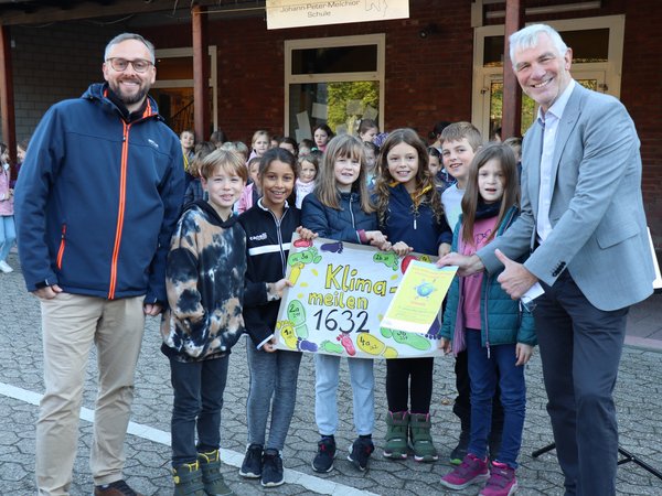 Zwei Männer und mehrere Kinder mit Plakat und Urkunde