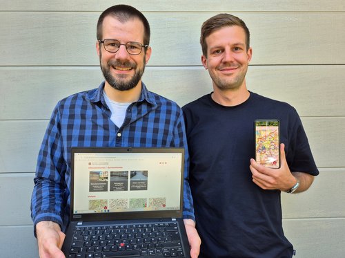 Zwei Männer nebeneinander - einer mit offenem Laptop, einer mit Handy 