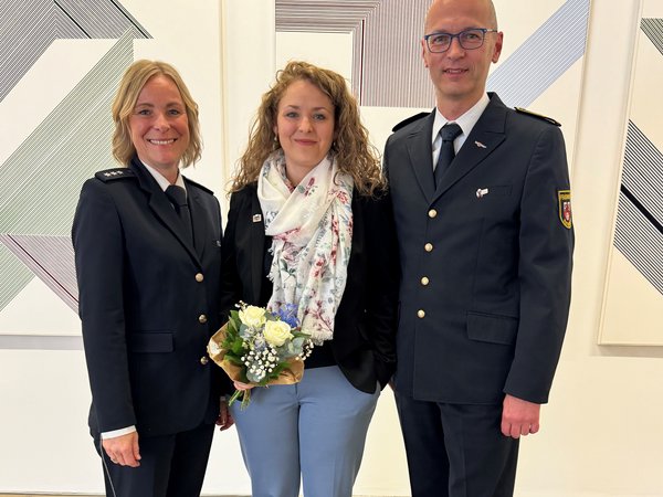 Frau mit Blumenstrauß in der Mitte, links und rechts Frau bzw. Mann mit Uniform