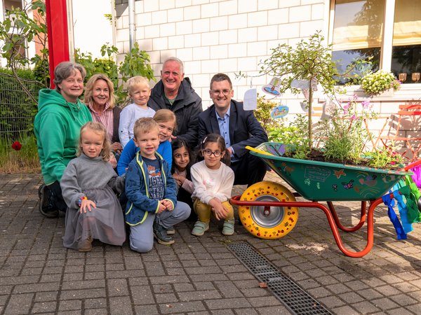 Mehrere Kinder und Erwachsene in der Hocke, daneben steht eine bepflanzte Schubkarre