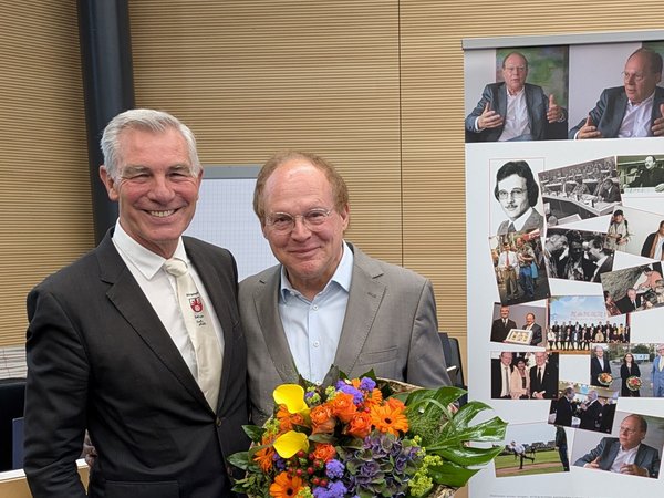 Zwei Männer, einer mit Blumenstrauß, neben einem Rollup 