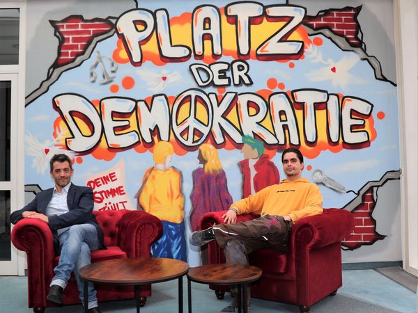 Zwei Männer sitzen in roten Sesseln vor einer Graffiti-Wand mit dem Schriftzug "Platz der Demokratie" 