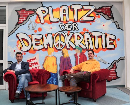 Zwei Männer sitzen in roten Sesseln vor einer Graffiti-Wand mit dem Schriftzug "Platz der Demokratie" 