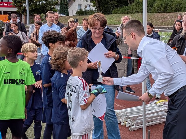 Ein Mann überreicht einem Jungen im Fußballtrikot einen Pokal, um sie herum stehen weitere junge Fußballspieler und Erwachsene. 
