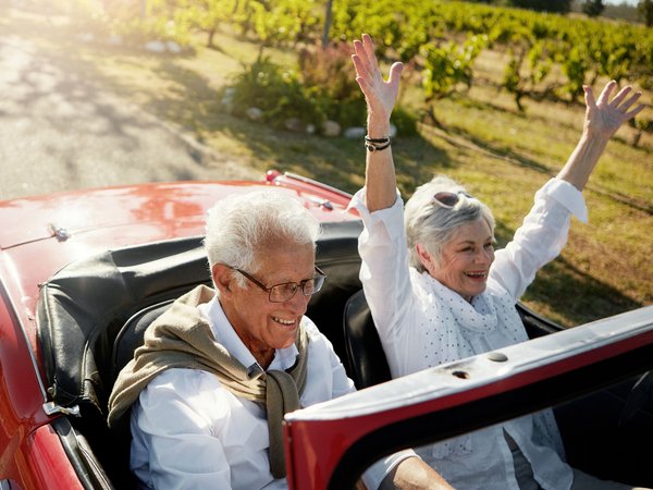 Ein älteres Paar sitzt lachend in einem roten Cabrio, die Frau hat beide Arme fröhlich in die Luft gestreckt.