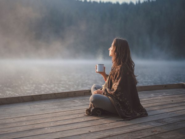Frau sitzt mit einer Tasse in der Hand auf einem Holzsteg an einem See.