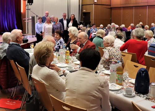 Viele ältere Menschen sitzen an Tischen in einem Saal und feiern zusammen