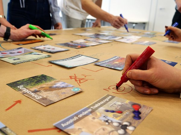 Karten und Stifte auf einem Tisch, drumherum agieren mehrere Personen