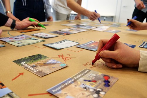 Karten und Stifte auf einem Tisch, drumherum agieren mehrere Personen