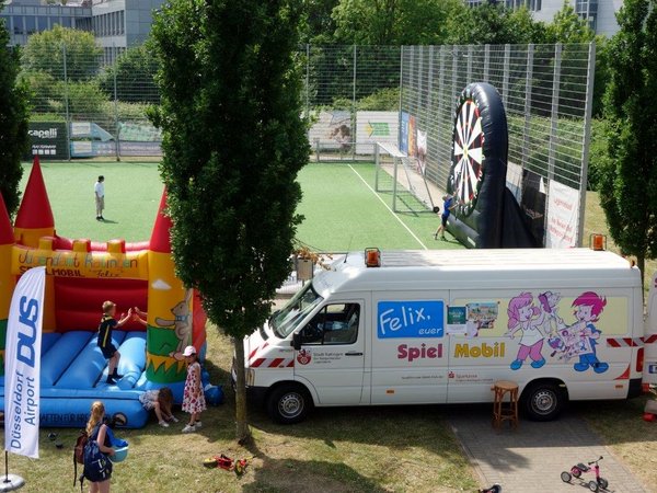 Auf einer Wiese stehen eine bunte Hüpfburg und ein weißer Spielmobil-Transporter mit Comicfiguren, dahinter eine übergroße Dartscheibe