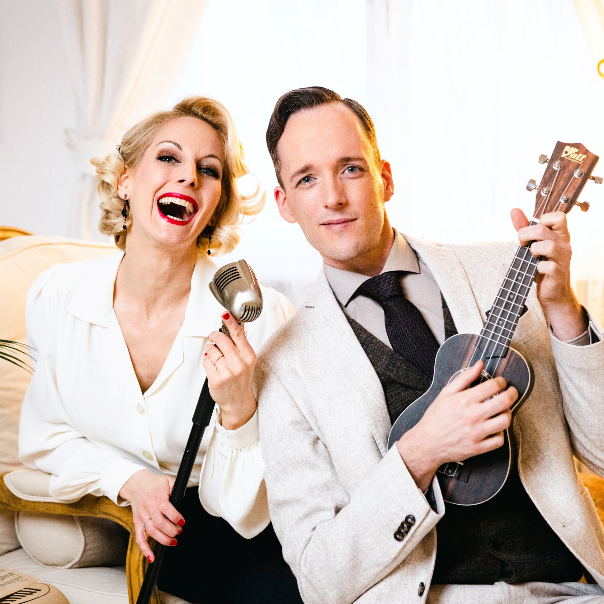 Die zwei Musiker sitzen im eleganten Outfit mit Instrument auf einem Sofa.