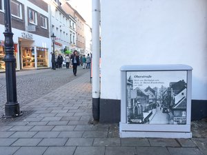 Das Foto zeigt einen Stromkasten in der Ratinger Innenstadt, welches mit einem historischen Foto der Oberstraße bedruckt ist.