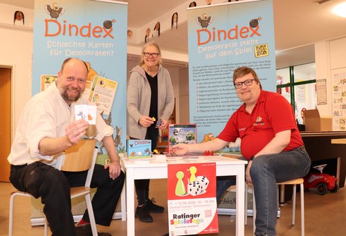 Zwei Männer sitzen an einem Spieltisch, einer hält Karten in den Händen. Eine Frau steht dahinter.