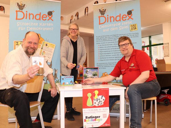 Zwei Männer sitzen an einem Spieltisch, einer hält Karten in den Händen. Eine Frau steht dahinter.