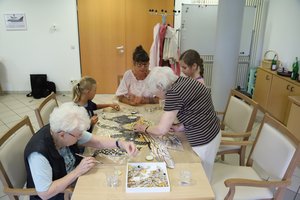 Tisch mit 3 Erwachsenen und 2 Kindern, auf dem Tisch liegt ein Mosaik-Bild an dem sie gemeinsam arbeiten