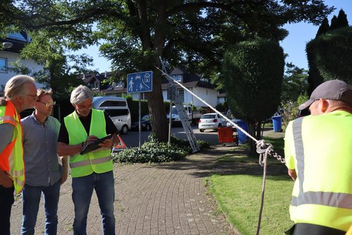 Ein Seil ist mit einem Baumstamm verbunden - ein Mann zieht am Seil, drei weitere schauen in Pläne.  