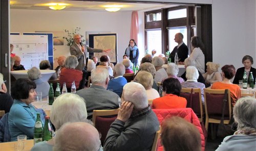 Bürgermeister mit Mikrofon spricht vor mehreren Personen, die an Tischen in einem Saal sitzen.