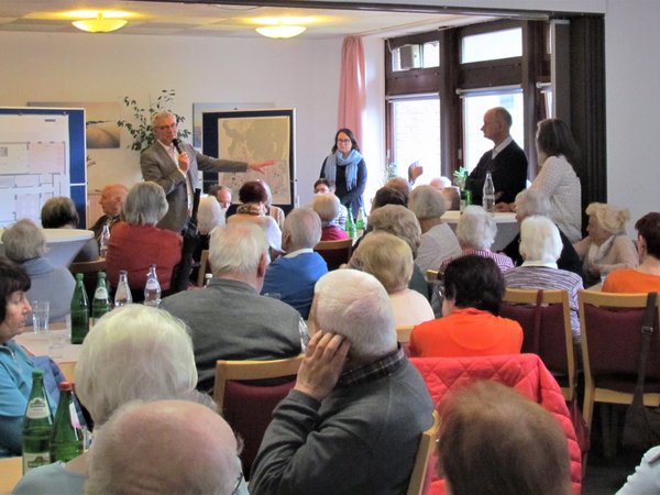Bürgermeister mit Mikrofon spricht vor mehreren Personen, die an Tischen in einem Saal sitzen.