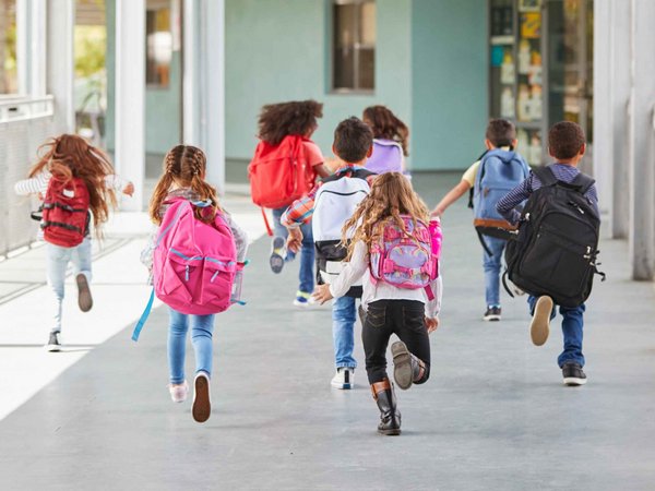 Sieben Kinder mit bunten Rucksäcken rennen über einen Schulgang.